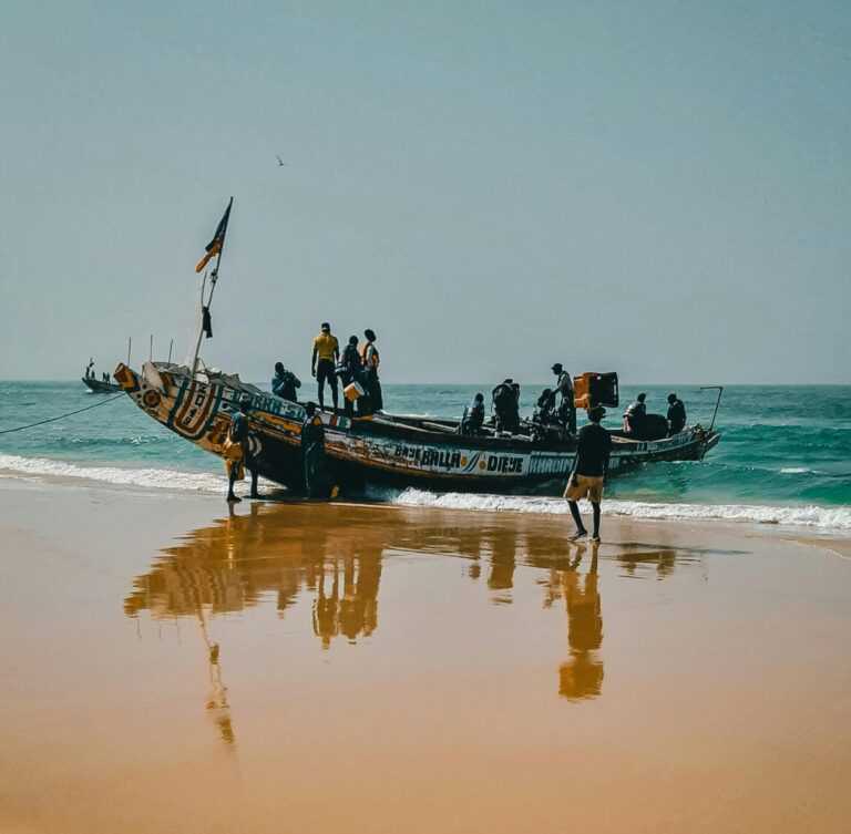 Colorful wooden fishing boat with crew on Kayar Beach, Senegal reflecting on sand under clear sky.