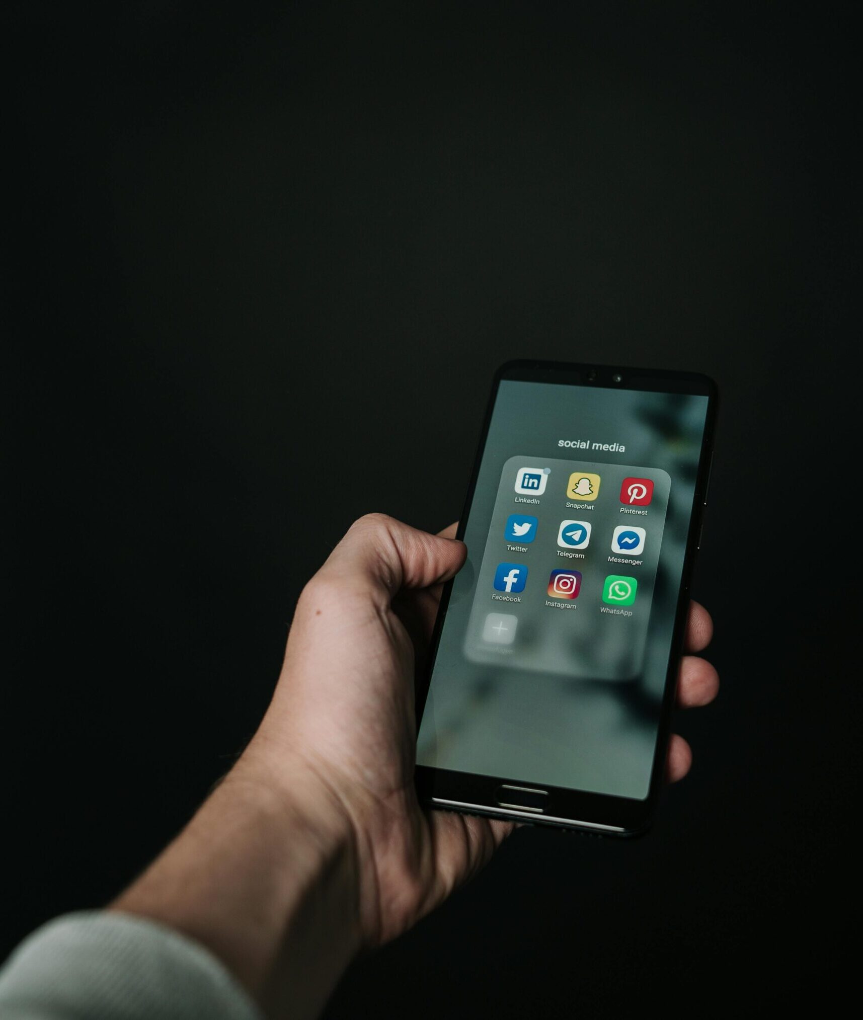 Close-up of a hand holding a smartphone displaying various social media app icons on a dark background.