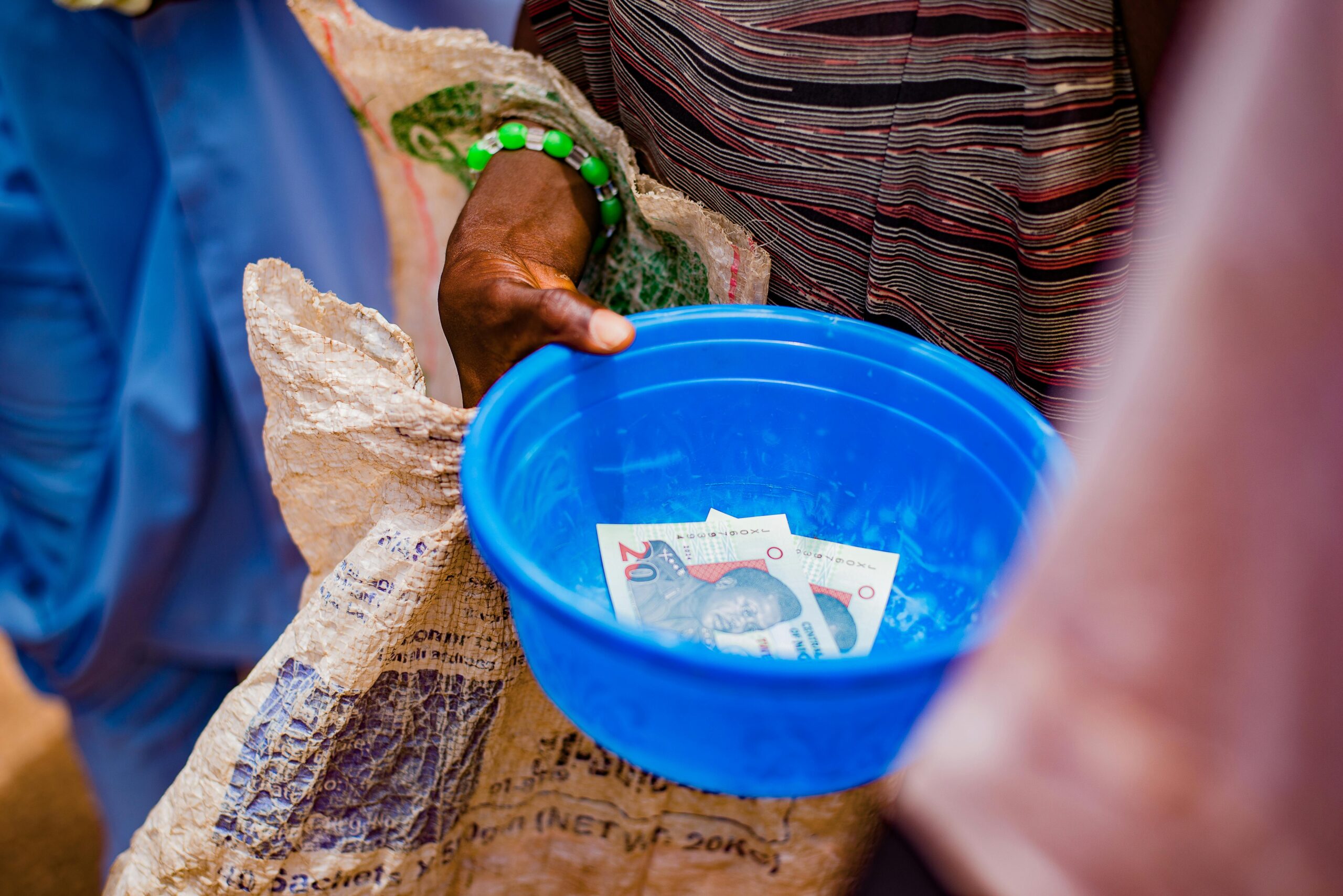 A close-up of a hand holding a blue bowl containing currency notes outdoors.