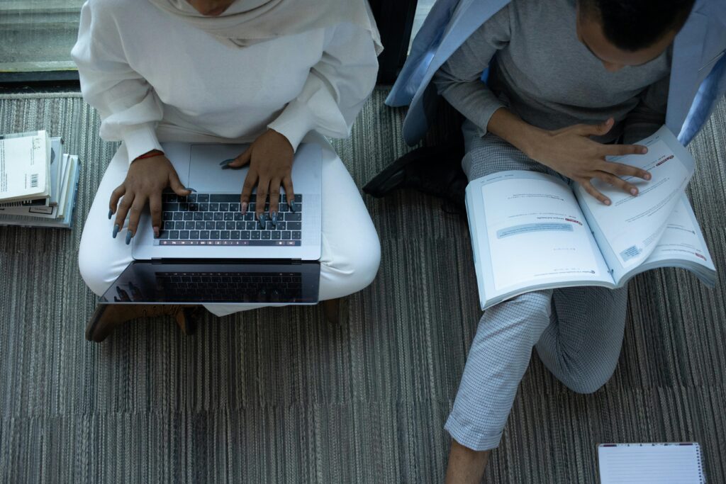 Two students studying with laptops and books in a library setting from above.