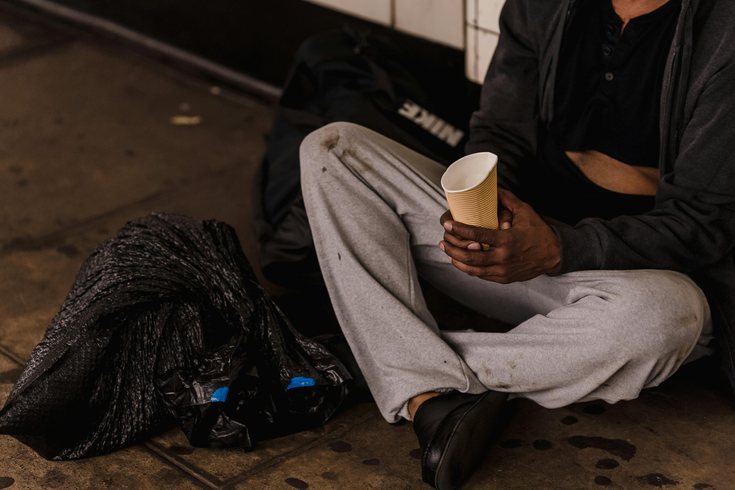 Close-up of a man sitting on a city street holding a paper cup, highlighting urban life challenges.