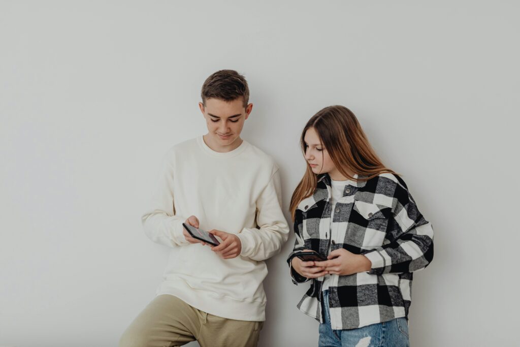 Teenage boy and girl using smartphones while leaning against a white wall indoors.