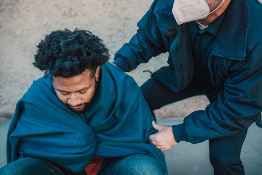 A paramedic assists an adult man covered with a blanket outdoors, showing care and support.