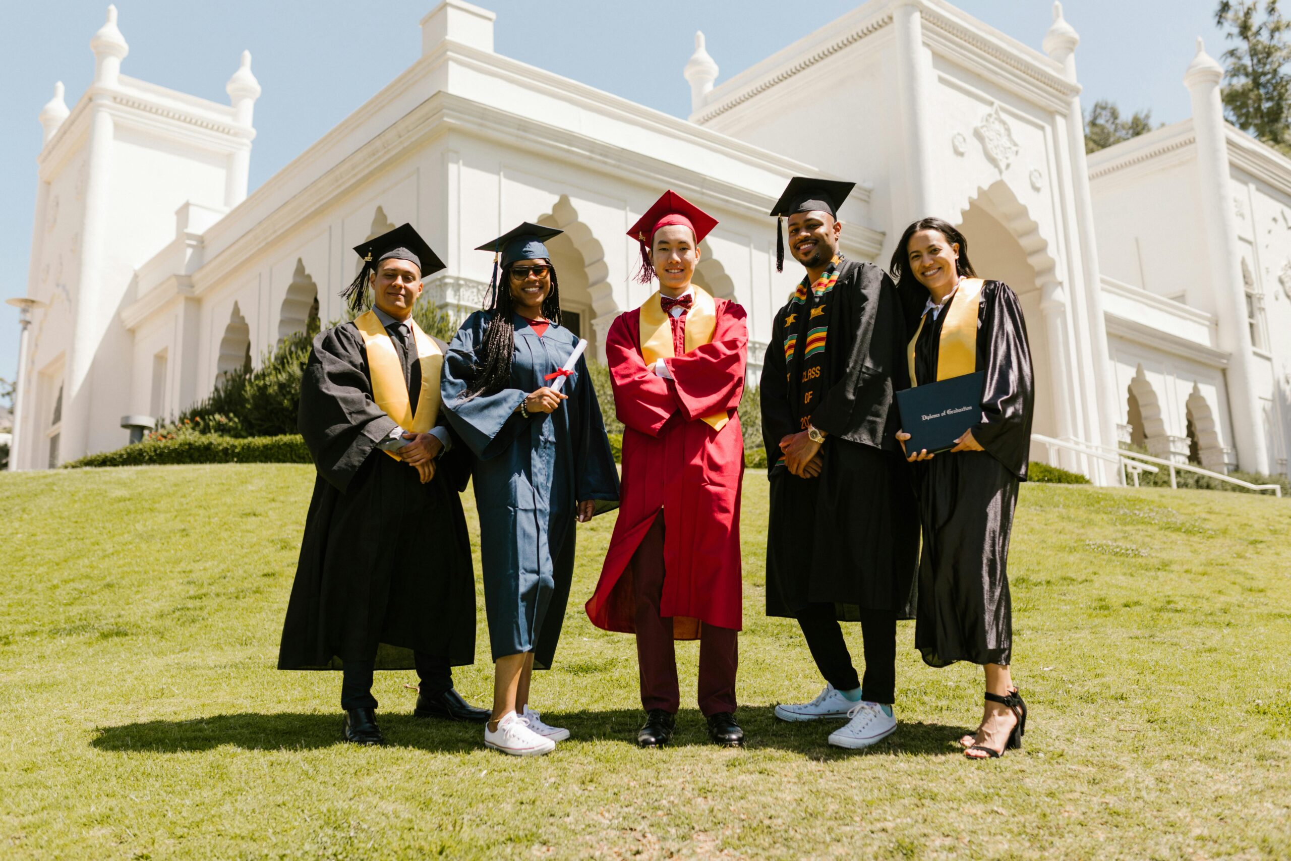 Five diverse graduates in caps and gowns posing happily outdoors at a university.