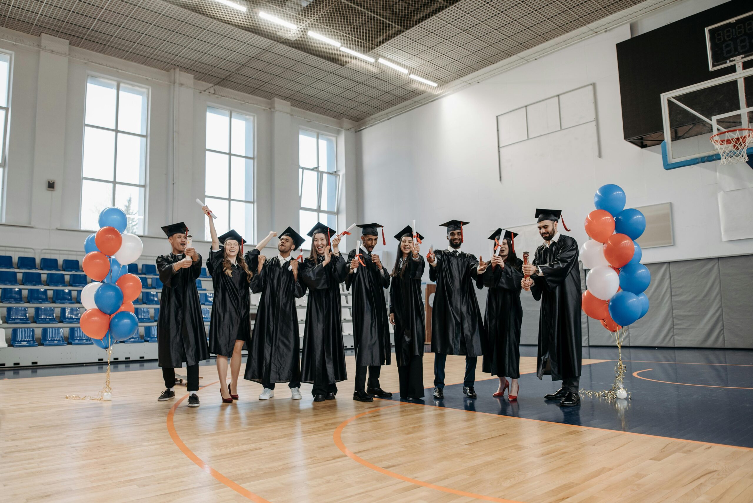 Group of happy graduates in gowns celebrating in an indoor sports hall with balloons.