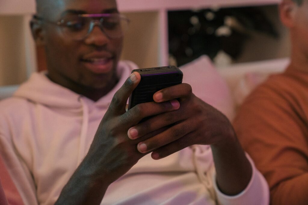 A man sitting indoors with friends, happily using his smartphone amidst warm lighting.
