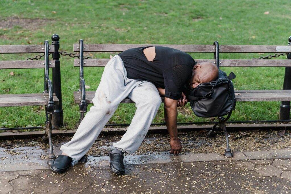 A man resting on a bench outdoors with a backpack, suggesting themes of homelessness and urban life.